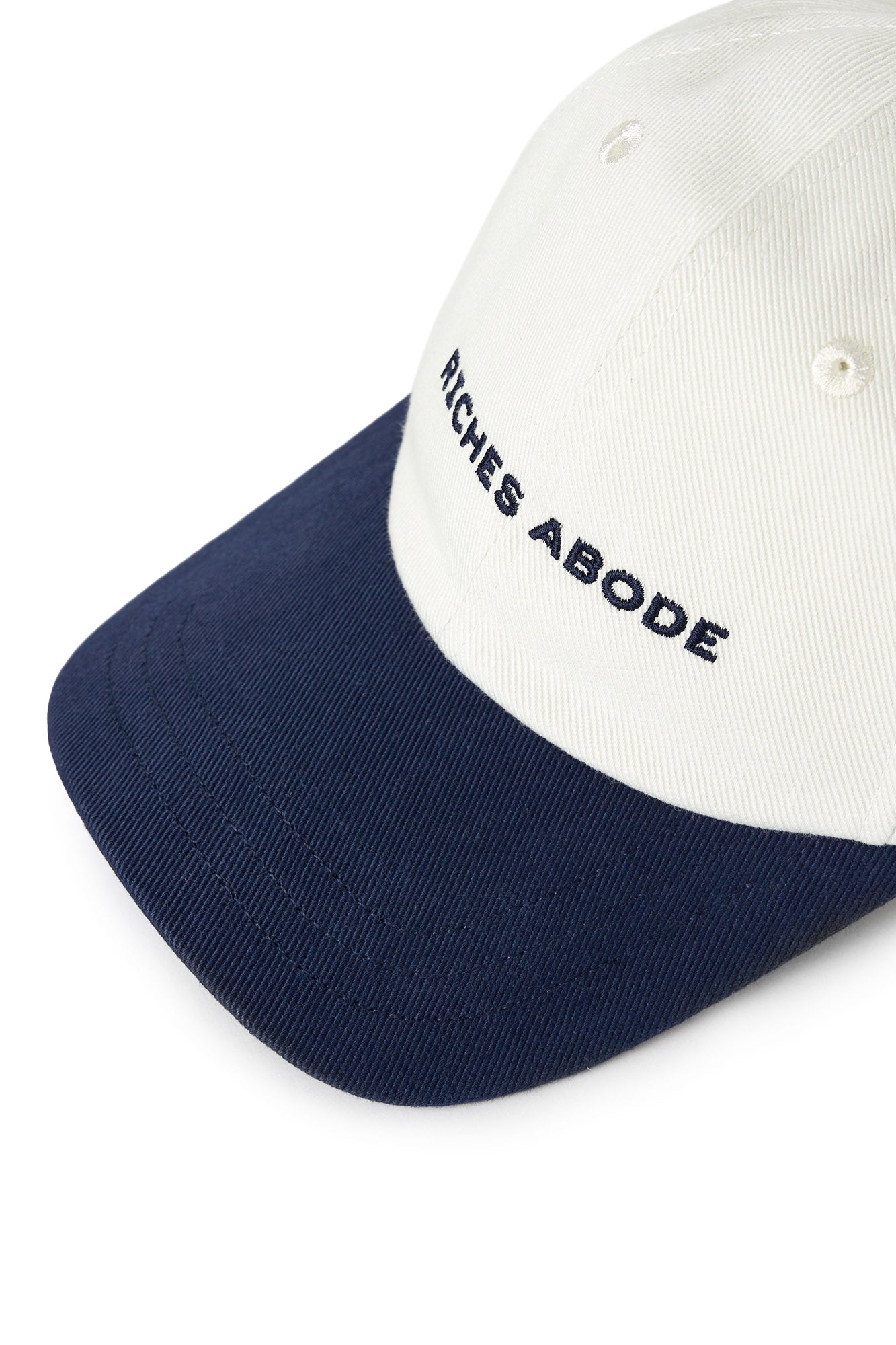 Riches Abode baseball cap in white with a navy brim, featuring an adjustable strap and eco-friendly construction. Ideal for versatile wear
