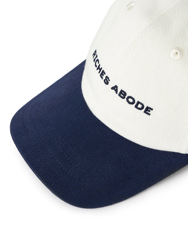 Riches Abode baseball cap in white with a navy brim, featuring an adjustable strap and eco-friendly construction. Ideal for versatile wear